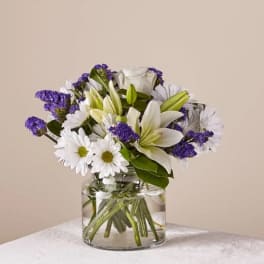 White lilies and daisies with purple filler in a clear glass vase