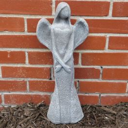 Tall gray stone angel statue with folded arms standing against a red brick wall.