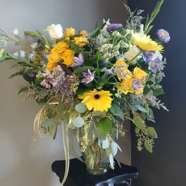 Mixed bouquet of yellow roses, gerbera daisies, and purple flowers in a glass vase