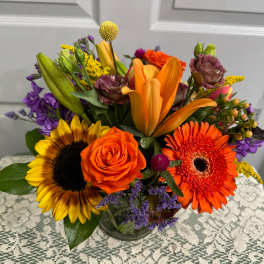 Mixed bouquet with sunflowers, orange roses, lilies, and gerbera daisies in a glass vase