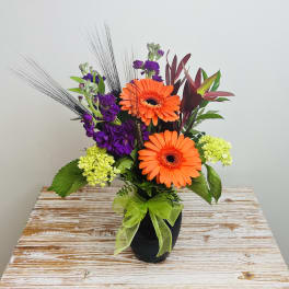 Orange gerbera daisies and purple flowers in a black vase with a green ribbon