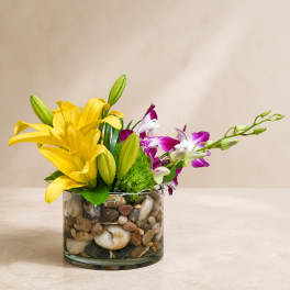 Yellow lilies and purple orchids in a pebble-filled glass vase