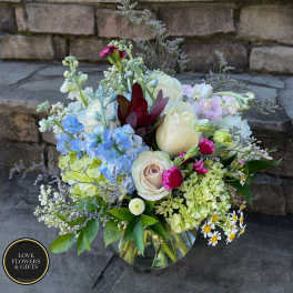 Mixed bouquet of cream roses, blue delphinium, green hydrangea and small daisies in a clear glass vase.