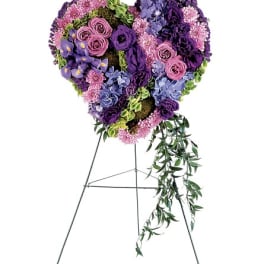 Heart-shaped floral standing spray in purple, pink, and blue with trailing greenery