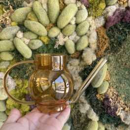 Amber glass watering can held in front of a moss wall
