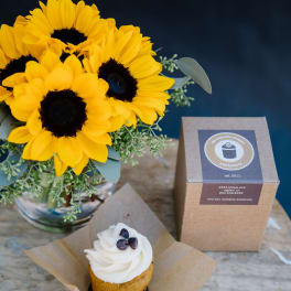 Sunflower bouquet in a glass vase beside a cupcake and a bakery box