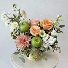 Peach roses and white blooms in a gold vase with green apples