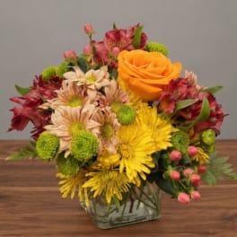 Mixed bouquet in a square glass vase with orange roses, yellow daisies, and red blooms