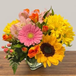 Mixed bouquet with pink gerbera daisies, yellow sunflowers, and orange roses in a glass vase
