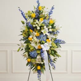 Standing floral spray with white lilies, blue flowers, and yellow roses on a white ribboned easel