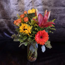 Mixed bouquet in a glass vase with orange roses, lilies, sunflowers, and a gerbera daisy