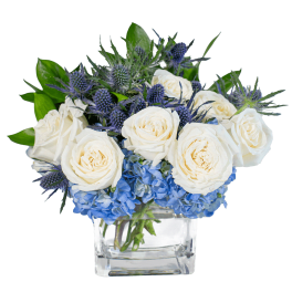 Low arrangement of white roses and blue hydrangeas in a clear glass cube vase