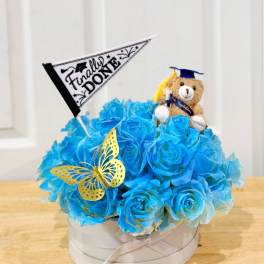 Blue rose arrangement in a round box with a teddy bear and graduation flag