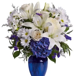 White lilies and daisies in a blue glass vase with a blue hydrangea cluster