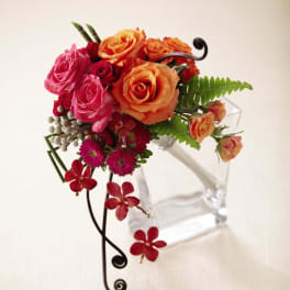 Modern arrangement of orange and pink roses with red accents in a tilted clear glass vase with metal scroll detail.