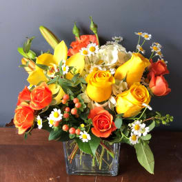 Bright arrangement of yellow roses, orange roses, lilies, and white daisies in a clear glass cube vase
