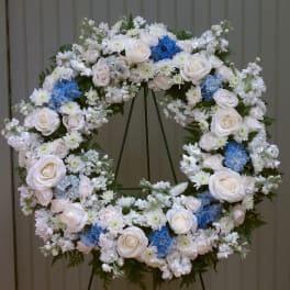 Large standing wreath of white roses and mixed white flowers with blue accents on a metal easel.