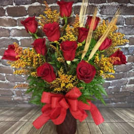 Red roses arranged in a tall vase with yellow filler and a red bow.