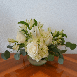 Low white arrangement of roses, lilies, and hydrangeas in a round glass vase