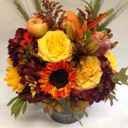 Mixed autumn bouquet with roses, sunflowers, apples, and lilies in a glass vase