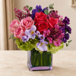 Mixed bouquet of pink, red, purple, and lavender flowers in a square glass vase