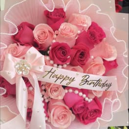 Round bouquet of hot pink and light pink roses with white wrap, pearls, and a Spanish birthday ribbon.