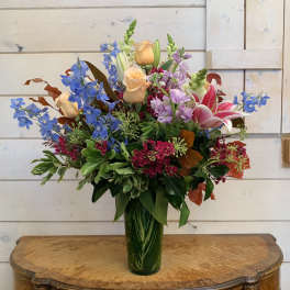 Mixed bouquet of blue delphinium, peach roses, pink lilies, and purple flowers in a green vase