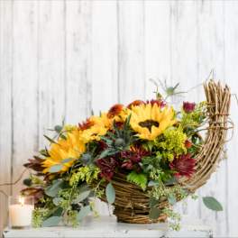 Basket arrangement with sunflowers and burgundy flowers beside a lit candle