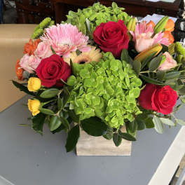 Mixed bouquet with red roses, pink blooms, and green hydrangeas in a square vase