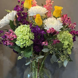 Mixed bouquet of yellow roses, white mums, and pink and green blooms in a clear glass vase
