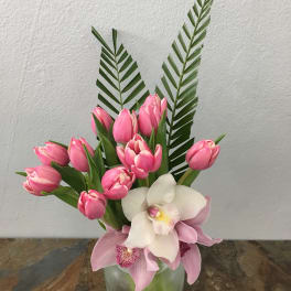 Pink tulips and orchids arranged in a glass vase with tall palm fronds.
