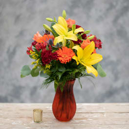 Mixed bouquet with yellow lilies, orange gerbera daisies, and red roses in a red vase