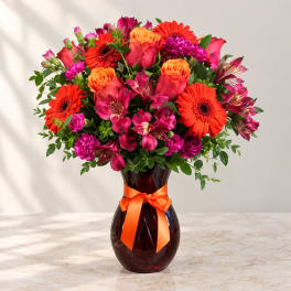 Mixed bouquet of orange, pink, and magenta flowers in a dark glass vase with ribbon