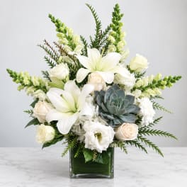 White lilies and roses arranged in a square glass vase with a succulent accent