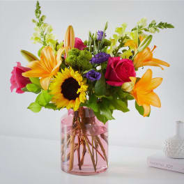 Colorful bouquet of roses, lilies, sunflowers, and purple blooms in a pink glass vase