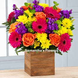 Bright mixed bouquet in a wooden cube vase with pink, orange, yellow, and purple flowers.