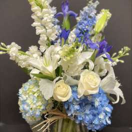 Tall arrangement of white roses, lilies, and blue hydrangeas in a clear glass vase