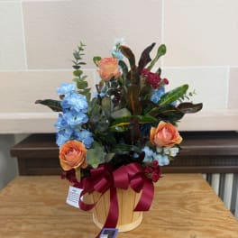 Peach roses and blue flowers arranged in a wooden basket with a burgundy ribbon.