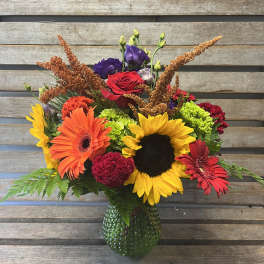 Colorful mixed bouquet with sunflowers, gerbera daisies, and roses in a green glass vase