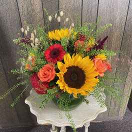 Low arrangement of sunflowers, orange roses, and red blooms in a green bowl on a small table