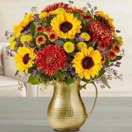Yellow sunflowers and orange mums arranged in a hammered gold metal pitcher