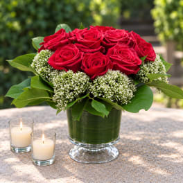Red roses arranged in a green glass vase with two lit candles nearby