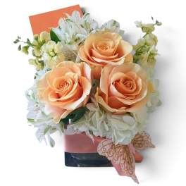 Peach roses and white flowers arranged in a square vase with a pink ribbon