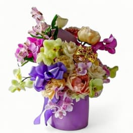 Mixed bouquet of pastel orchids and roses in a purple vase with bow and small gold crown decoration