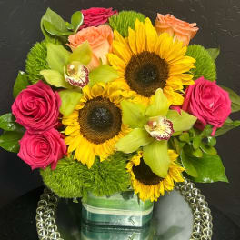 Bright bouquet of sunflowers, pink roses, and green orchids in a glass vase.