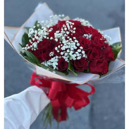 Handheld bouquet of red roses with white filler flowers wrapped in paper and tied with a large red bow.