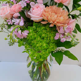 Tall glass vase of pink roses, carnations, lisianthus, peach mum, and green hydrangea.