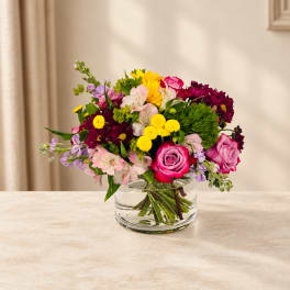 Mixed bouquet of pink roses, yellow blooms, and purple flowers in a clear glass vase