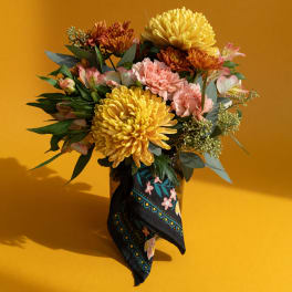 Bouquet of yellow, pink, and orange flowers in a dark vase with a patterned scarf
