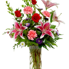 Tall glass vase with red roses, pink lilies, pink carnations, and purple accent flowers.
